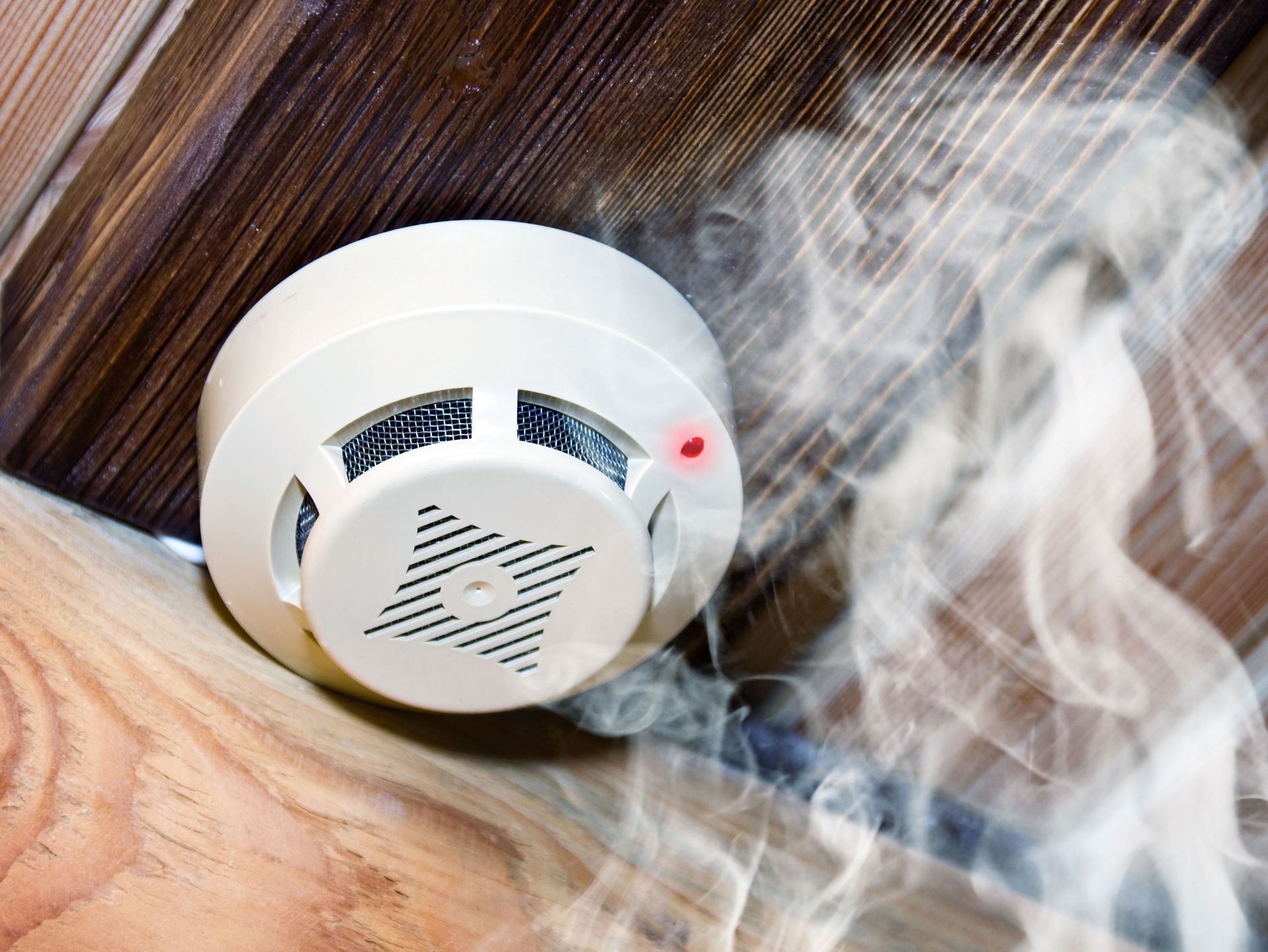 Smoke detector on a wooden ceiling with smoke billowing nearby and a red warning light on.