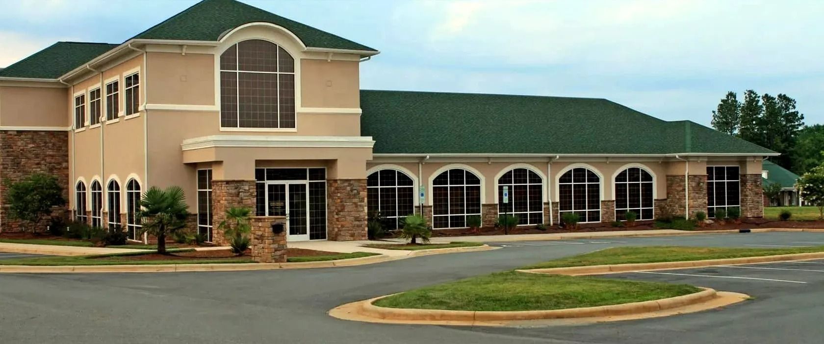 Two-story beige office building with green roof and arched windows beside a parking lot