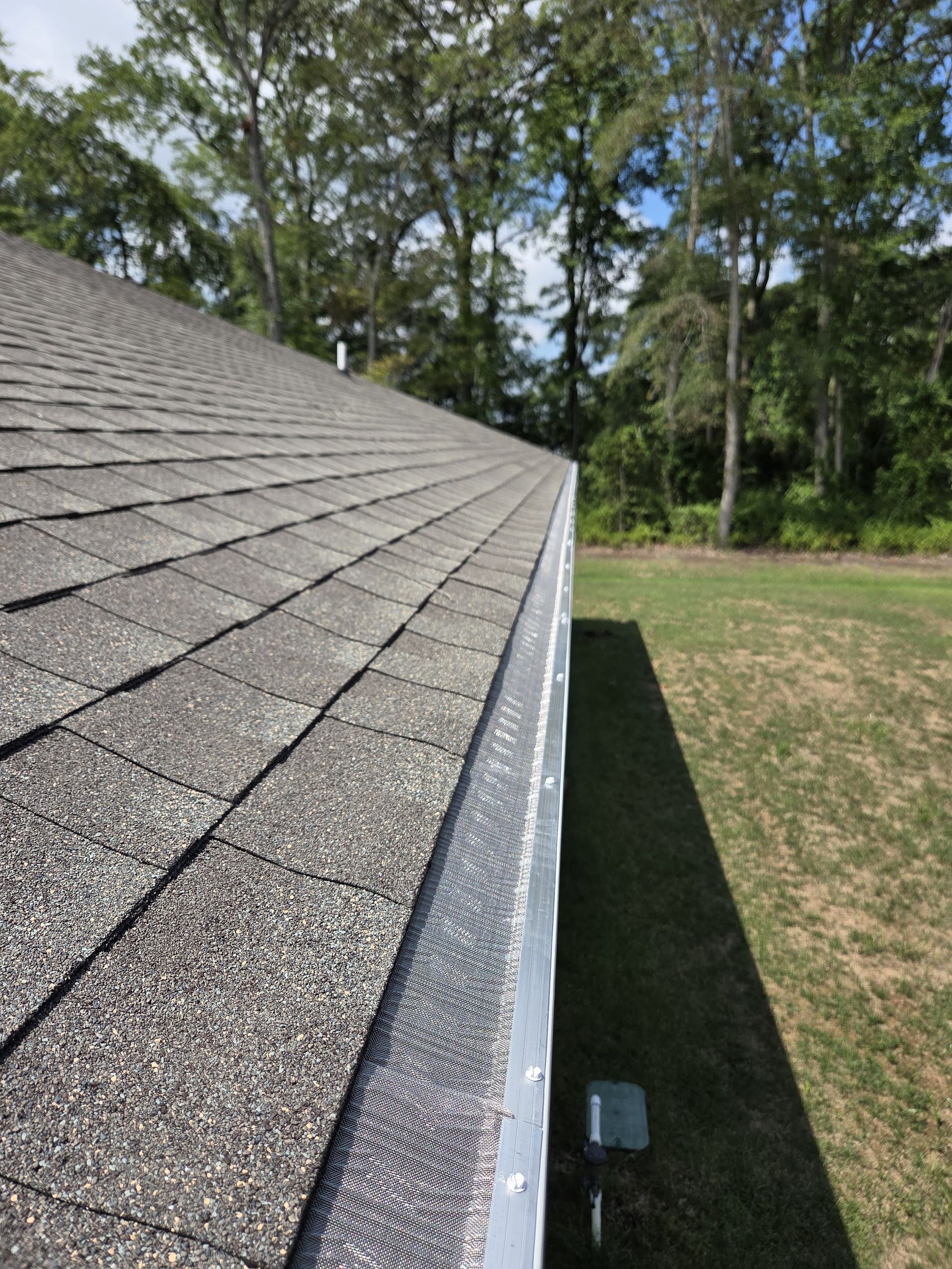 Roof shingles along a house edge beside a grassy yard and trees on a sunny day