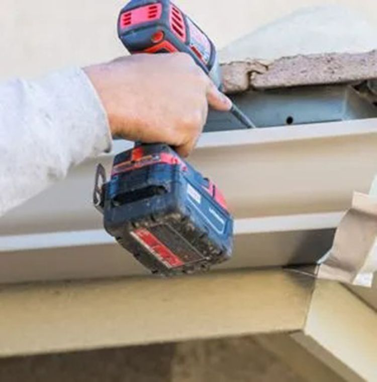 Hand holding a red and black power drill to a white soffit under a roof edge