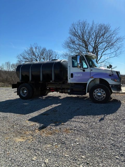 Purple dump truck parked on gravel under a clear blue sky, side view with leafless trees in back.