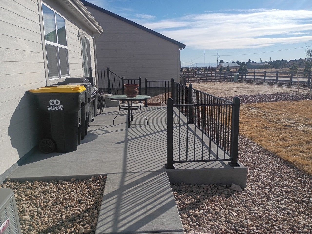 A concrete patio with a black railing, trash cans, and a table. Gravel yard and blue sky.