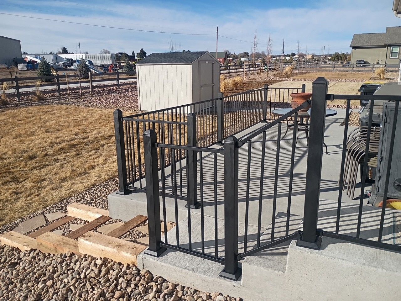 Black metal railing on a concrete patio with steps leading to a yard with a shed in the background.