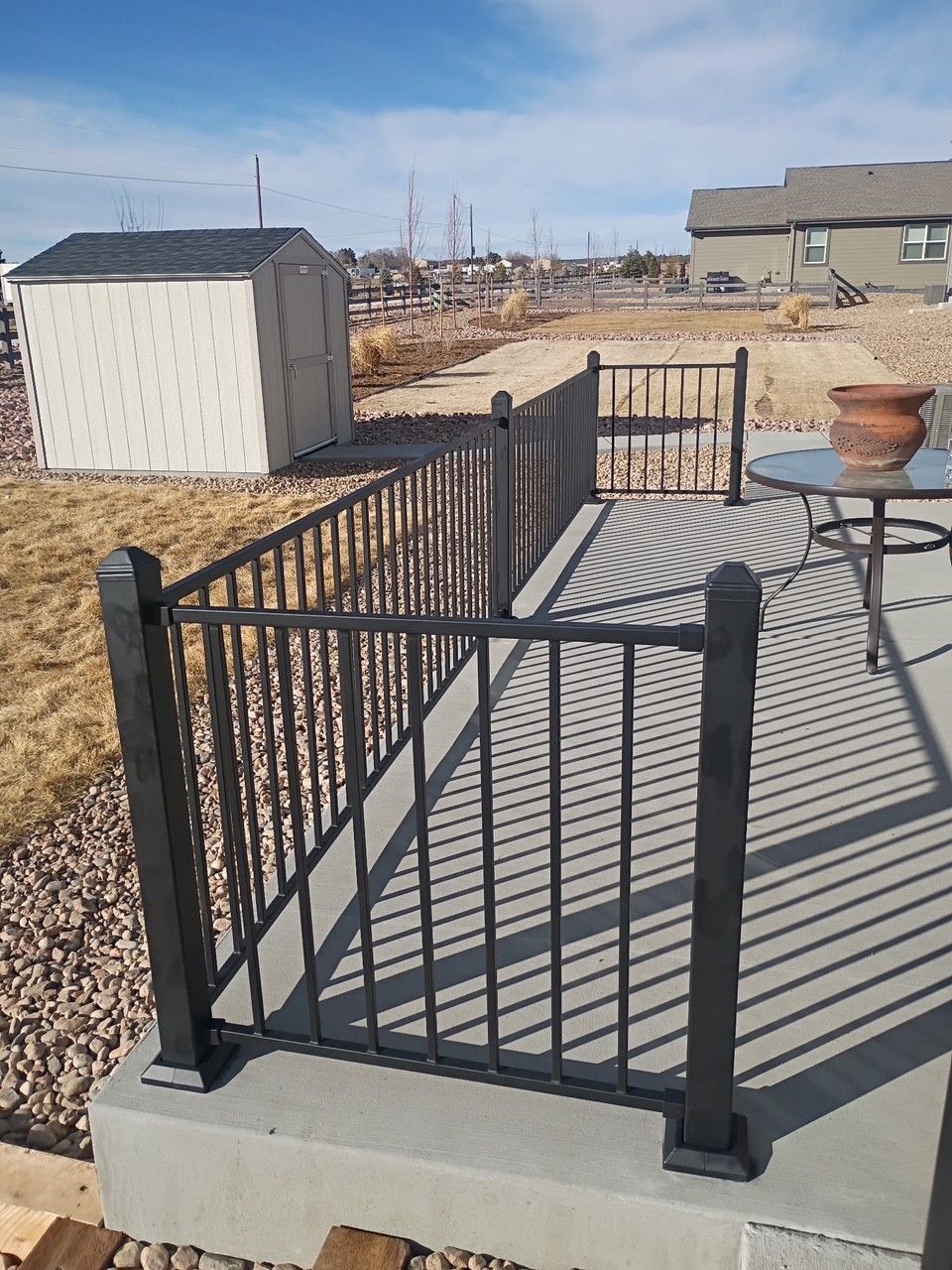 Black metal railing around a concrete patio with a shed and landscape in the background.