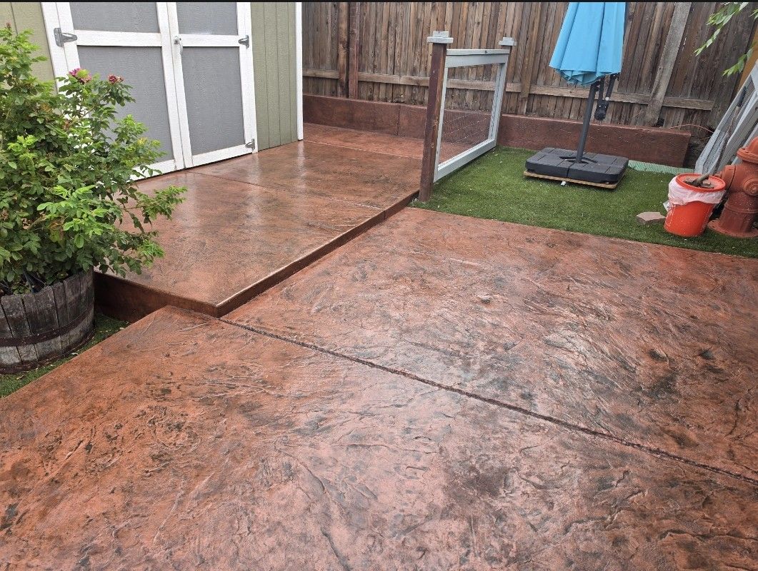 Wet concrete patio beside a fenced yard with a small grass area and outdoor furniture stand