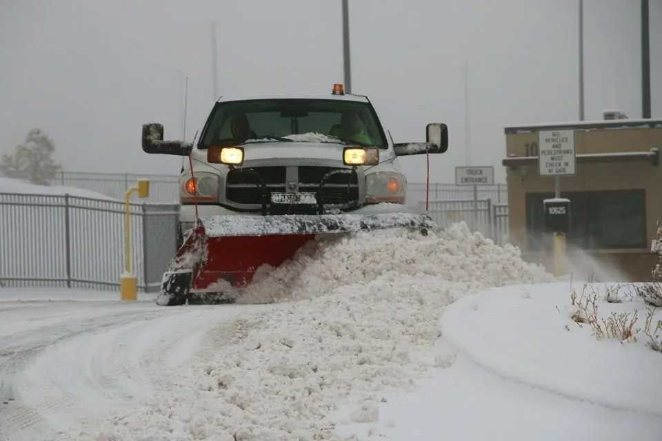 Snowplow clearing a snow-covered road in front of a gate during a snowstorm.