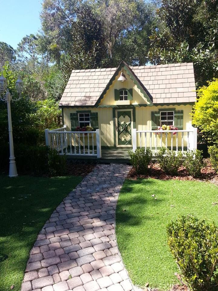 Yellow playhouse with green trim, a brick path, and surrounding greenery.