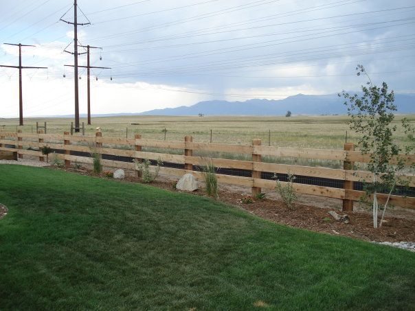 Green grass lawn with wooden fence, grassy field, power lines, and distant mountains under cloudy sky.