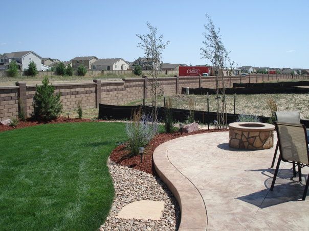Lush green lawn with landscaped beds, patio with fire pit, and decorative wall in a sunny backyard.
