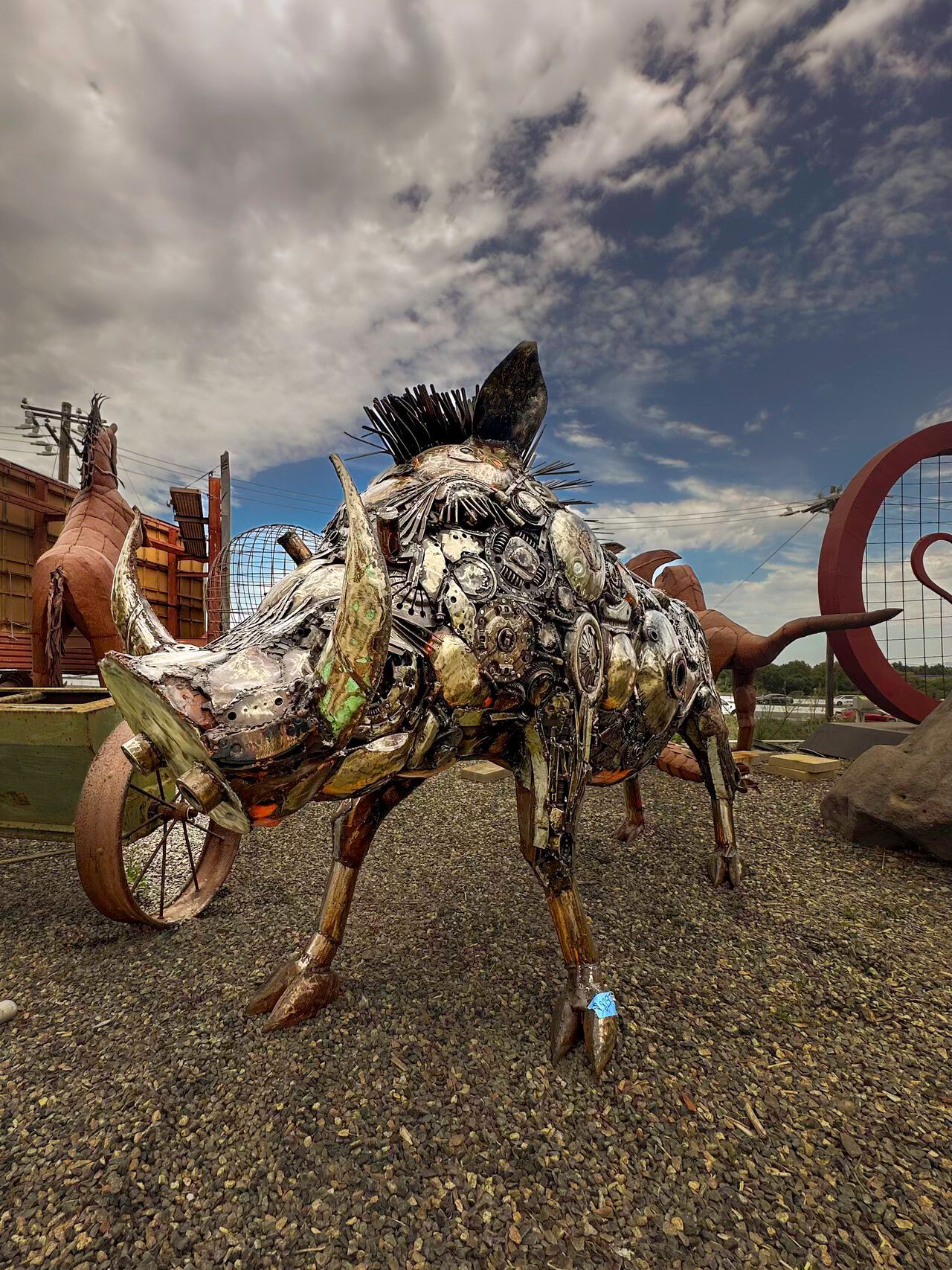 A statue of a boar made out of junk is standing on a gravel road.