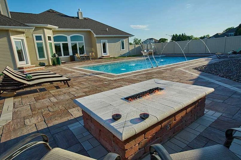 Backyard with pool, fire pit, lounge chairs, and house. Blue sky, brick and stone patio.