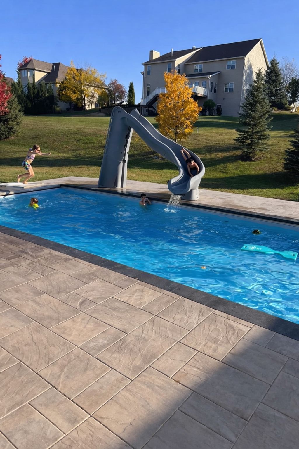 A rectangular pool with a water slide. Children play in the water. The setting is a backyard with houses in the background.
