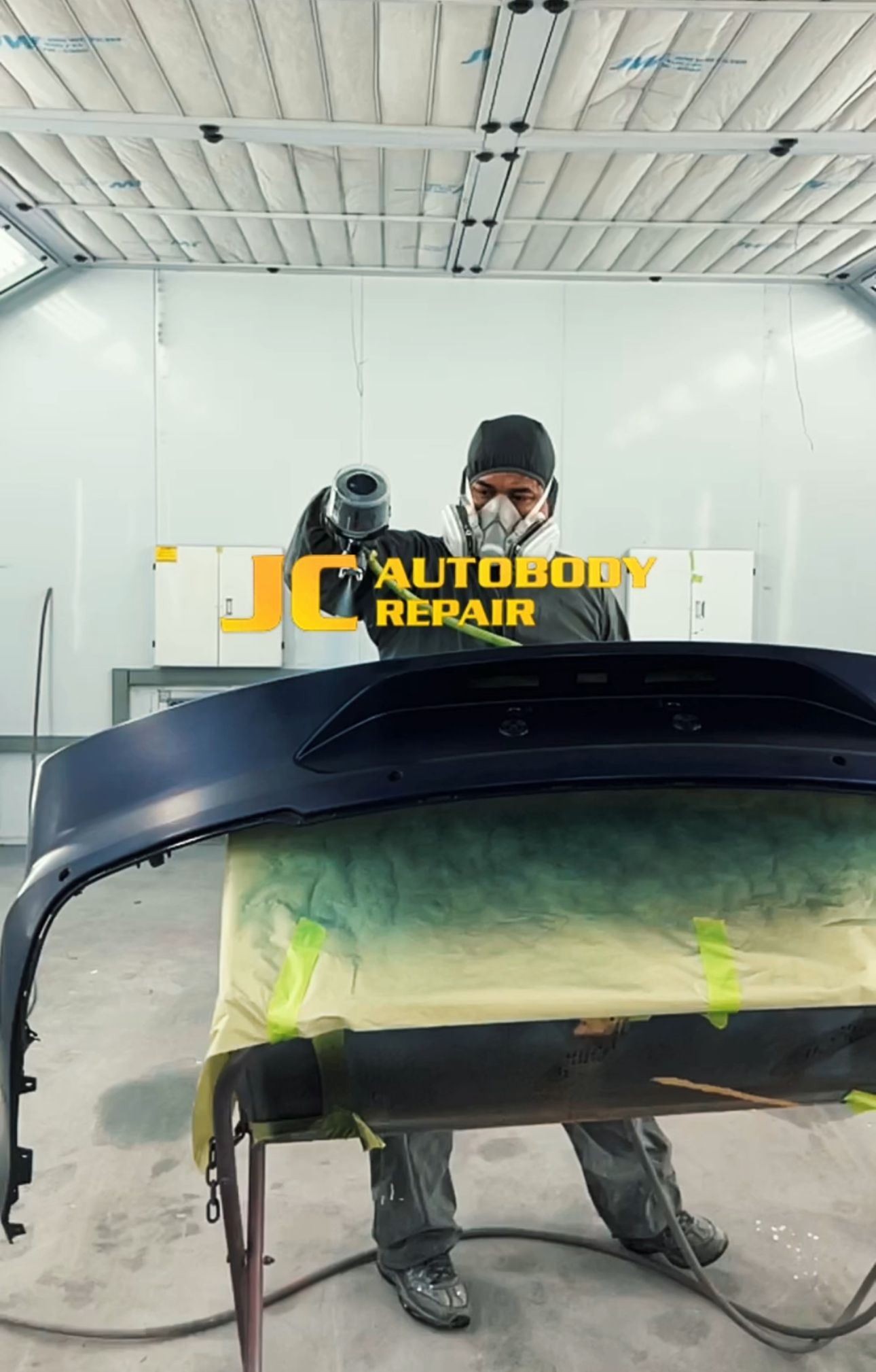 A man is painting a car bumper in a paint booth.