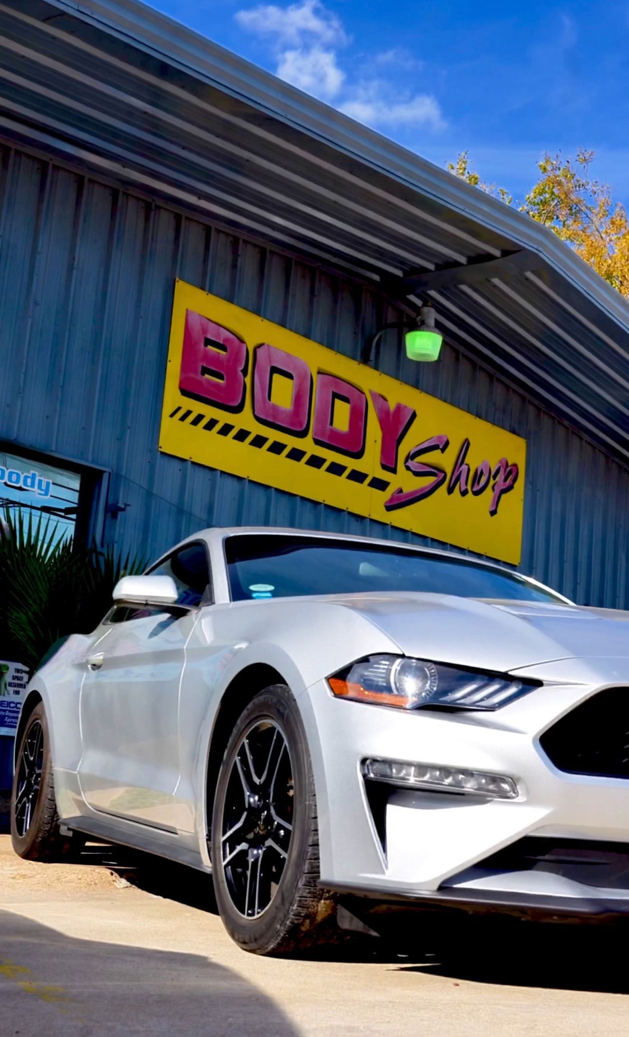 A white mustang is parked in front of a body shop.