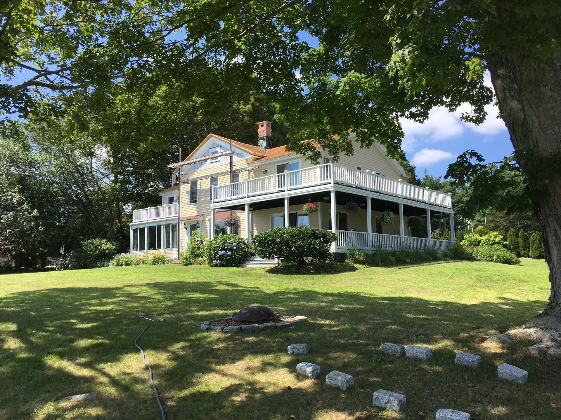Large white farmhouse with wraparound porch on a sunny lawn under leafy trees