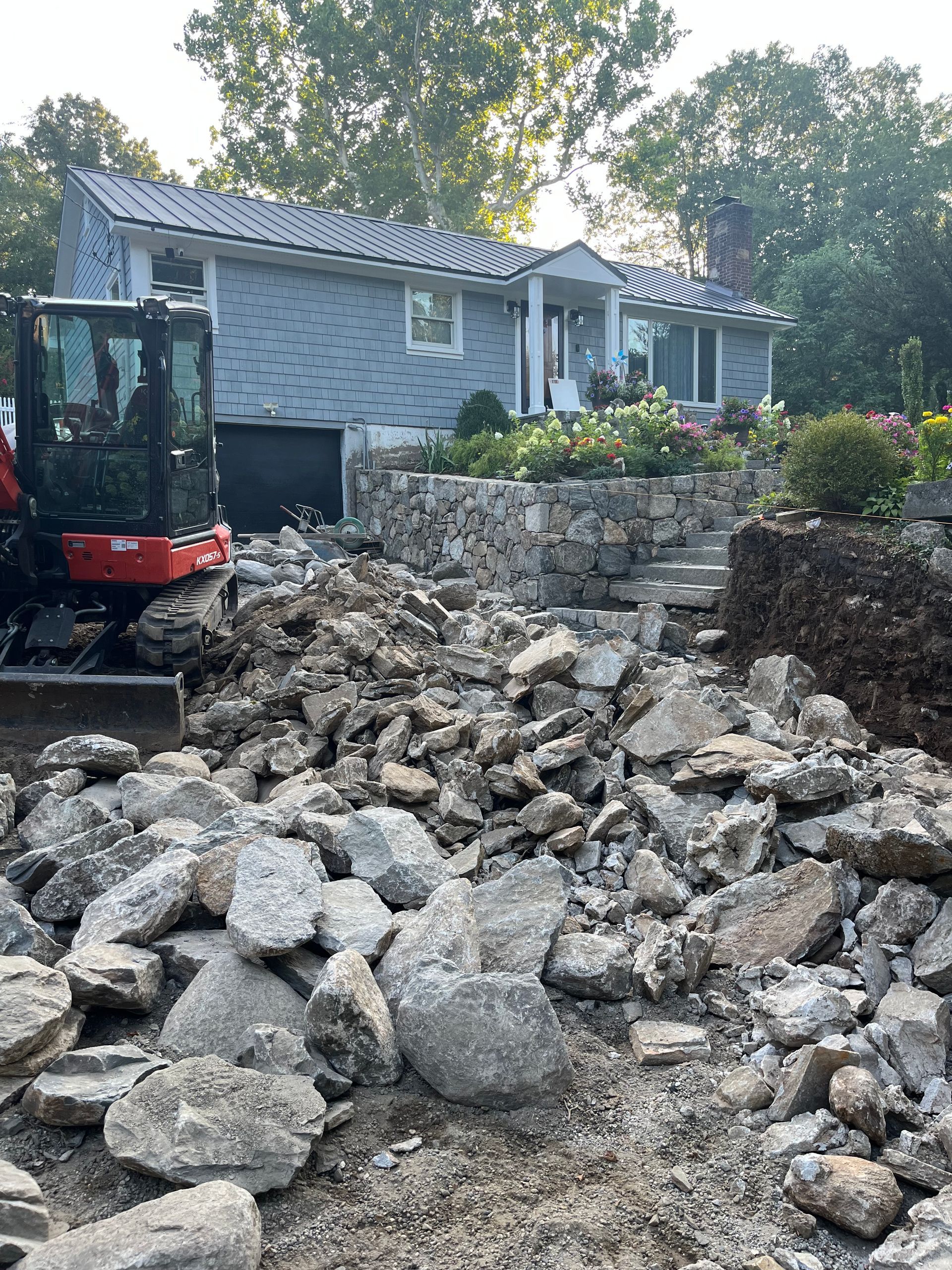 Excavation site at a light blue house. Pile of rocks and excavator, stone retaining wall.