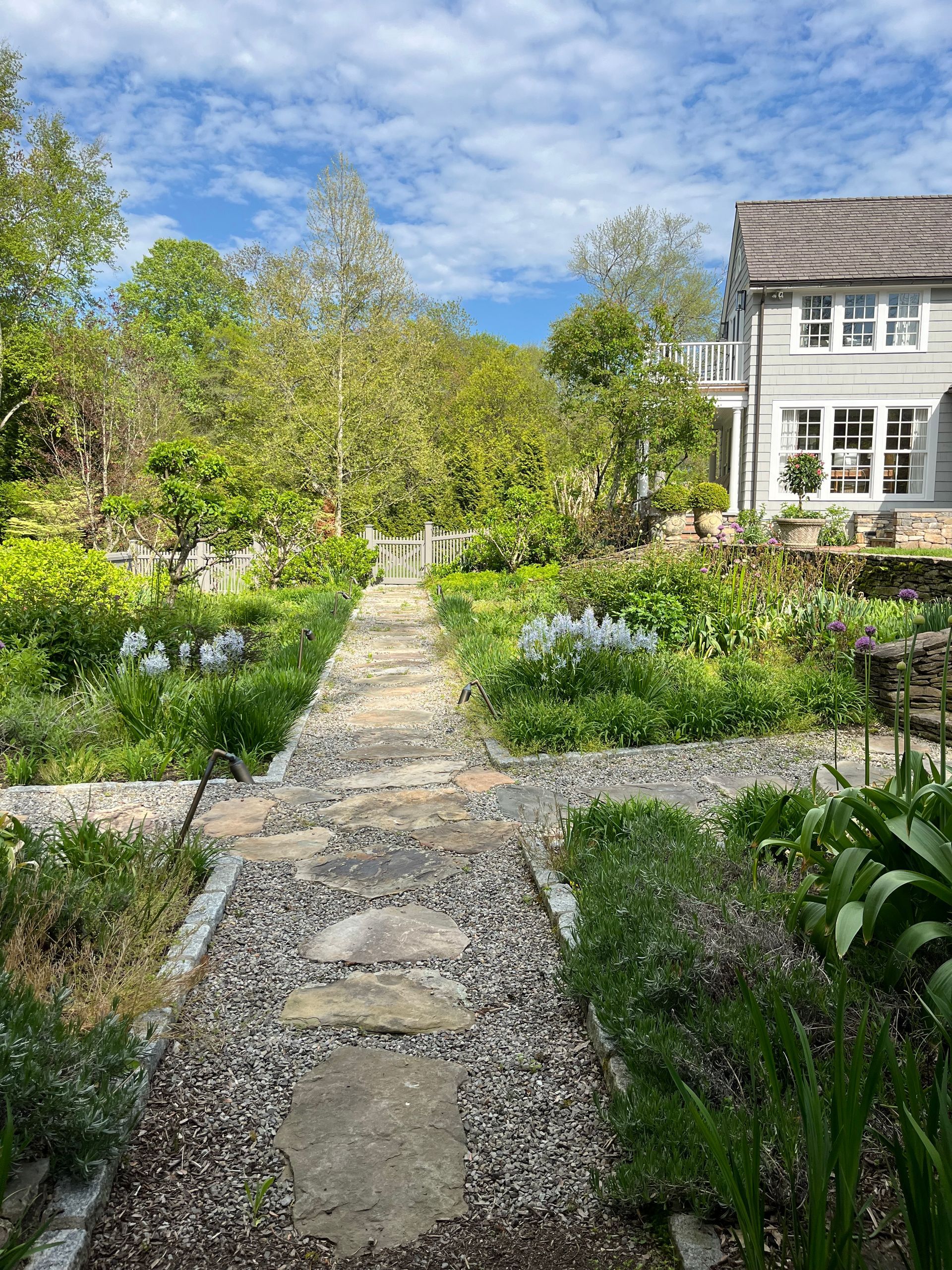 Stone path leads through a lush garden toward a gray house under a bright blue sky.