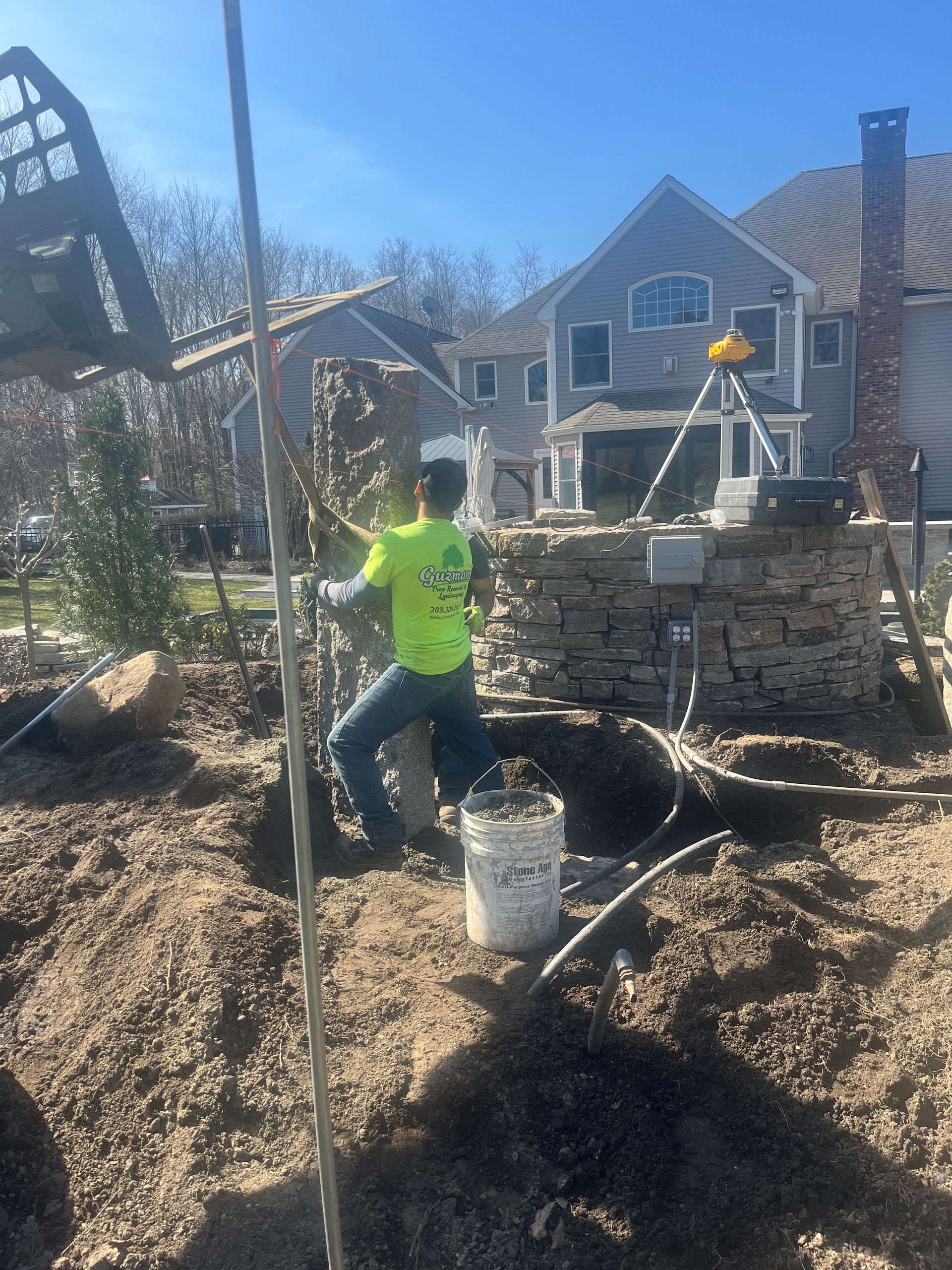 Man working on stone wall construction; digging in the soil. A luxury house is in the background.