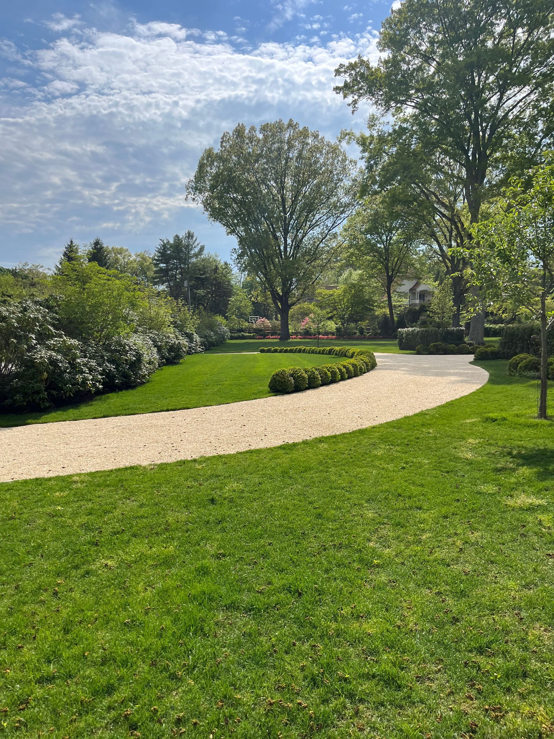 Grassy path curves through a lush green park with trees and shrubs under a partly cloudy sky.