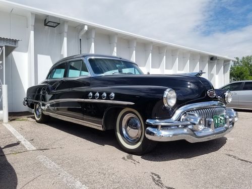 Black 1950s Buick Roadmaster with chrome trim and white wall tires parked outside a white building.