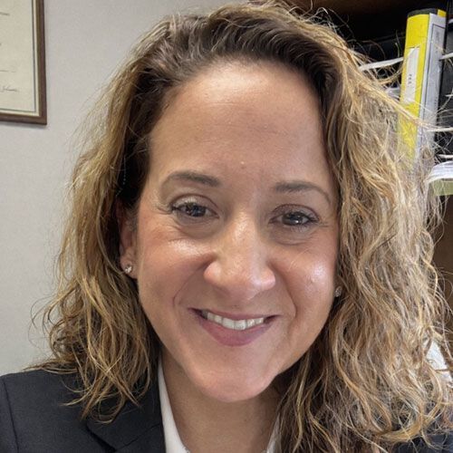 Woman in a suit smiling at the camera, with curly light brown hair, and a neutral background.