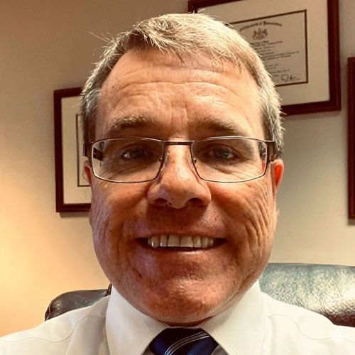 Man with glasses and tie, smiling, indoors in front of framed documents.