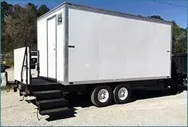 White mobile trailer with black steps and twin axles parked outdoors on gravel