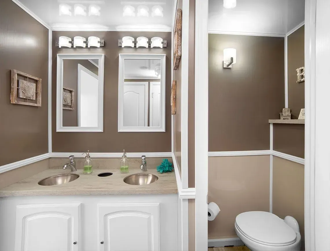Bathroom with double vanity, mirrors, tan walls, and a separate toilet area with white wainscoting.