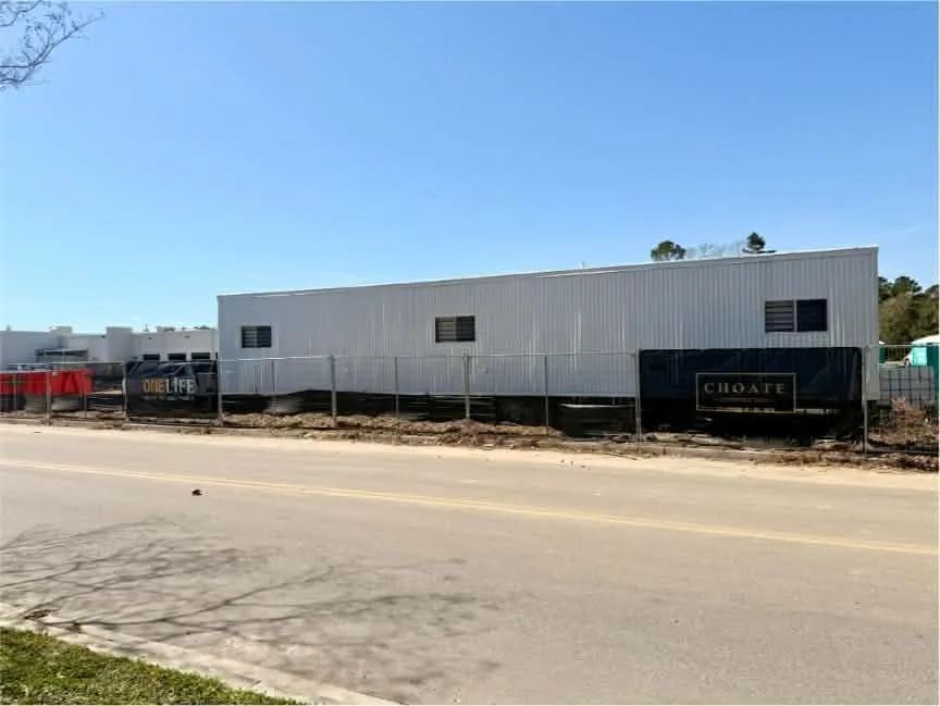 White construction trailer beside a road under a clear blue sky with parked equipment nearby