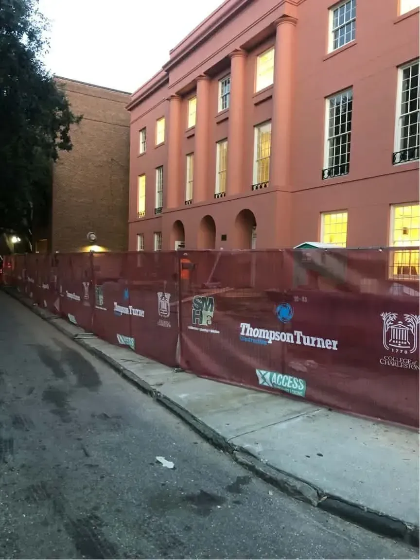 Red brick building behind a maroon construction fence along a narrow street at dusk
