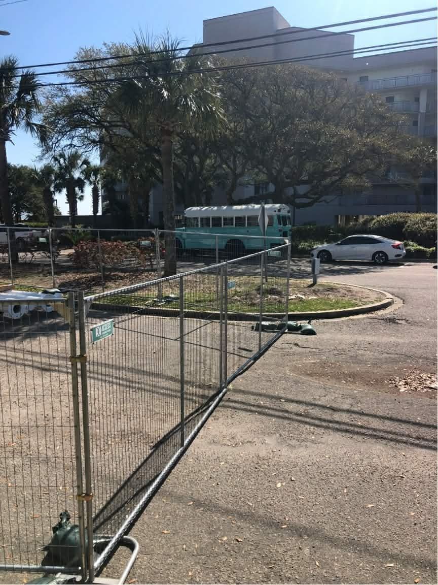 Temporary metal fencing along a roadside beside a tree and parked cars near a building.