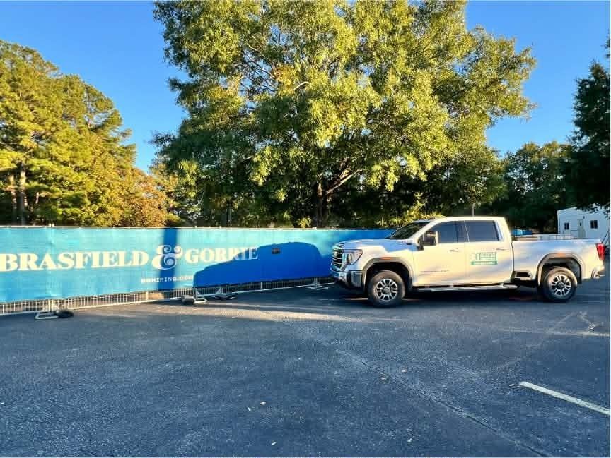 White pickup truck parked by a blue Brasfield & Gorrie construction fence under trees.