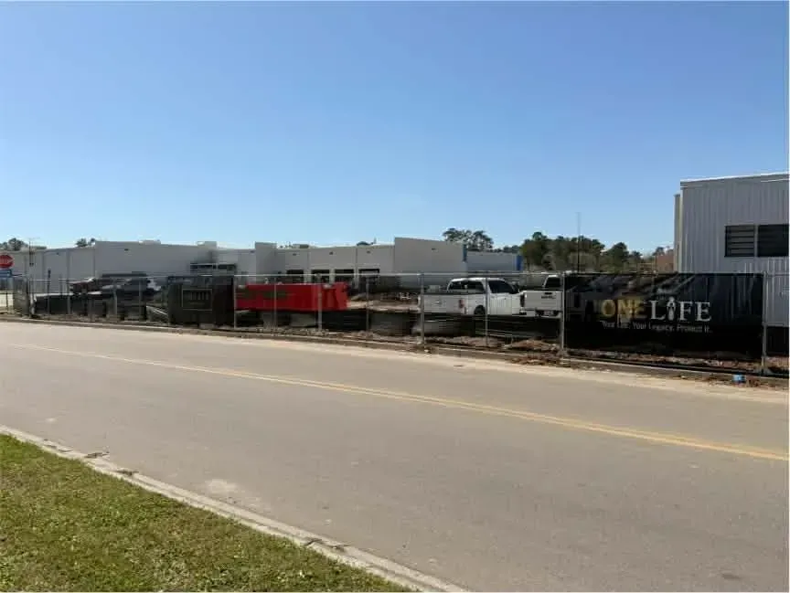 Roadside view of a fenced commercial lot with parked trucks, buildings, and a OneLife sign under a clear sky