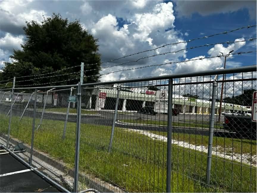 Chain-link fence beside a roadside parking lot under a blue sky with clouds, trees, and a low building in back