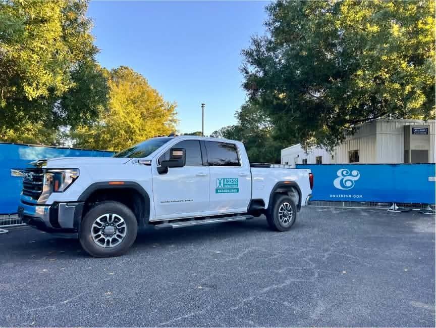White pickup truck parked beside a blue fence and building in a lot, with trees in the background