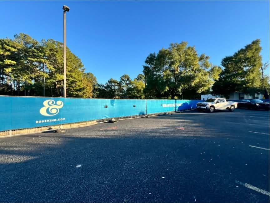 Outdoor parking lot with a blue fence, trees, and a few parked vehicles under a clear sky