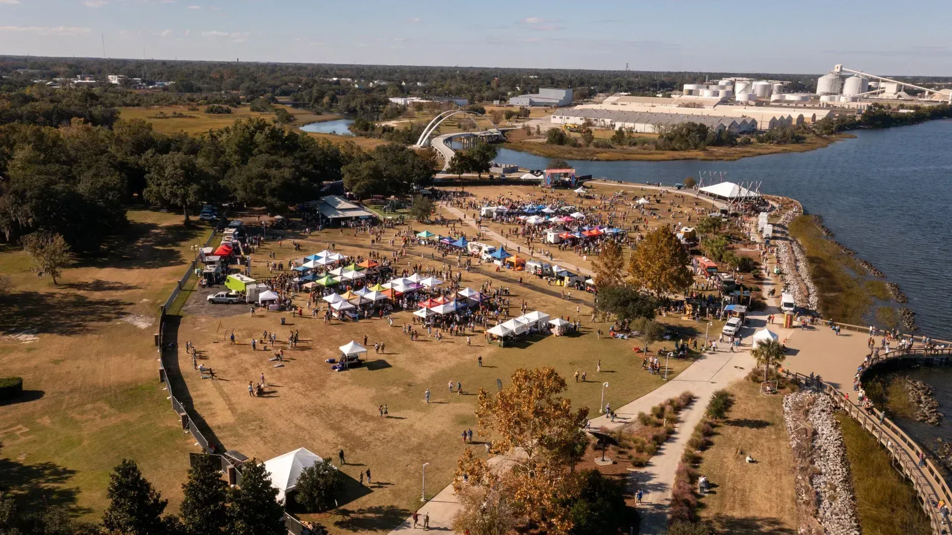 Aerial view of a crowded outdoor event by the water with tents, vehicles, and a shoreline path