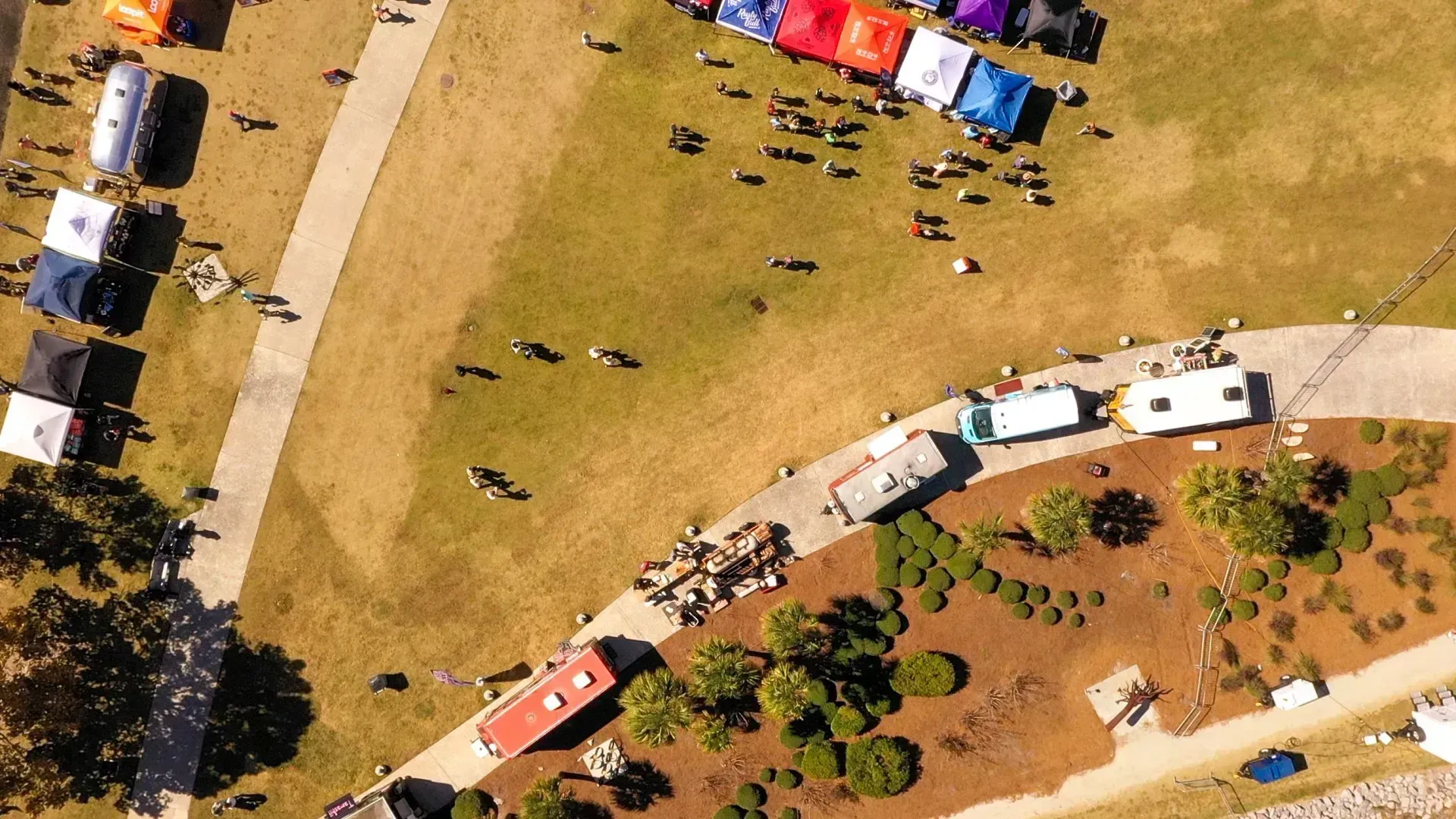 Aerial view of people gathered on a grassy area beside a curved road with parked cars and colorful tents.