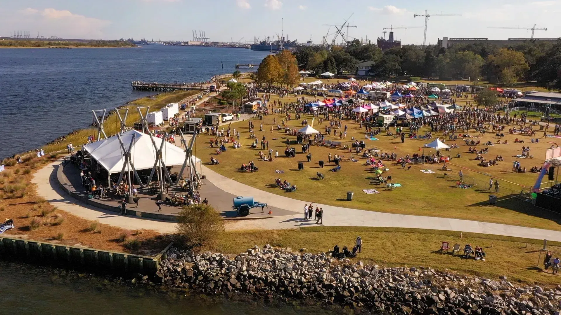 Crowded riverside park with tents, people on the grass, and water along a curving stone shoreline