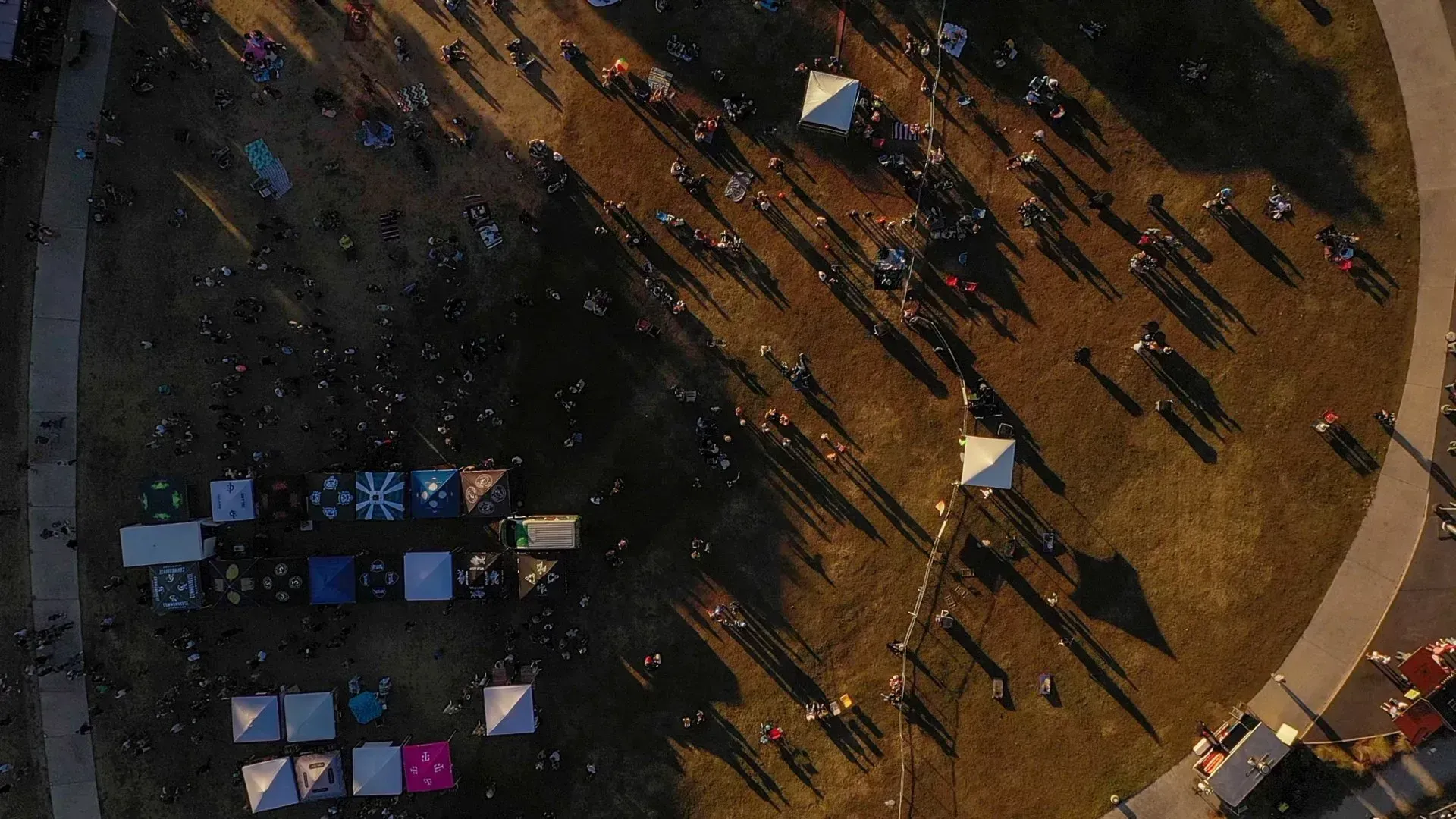 Aerial view of people casting long shadows in a plaza with colorful tents or tables nearby.