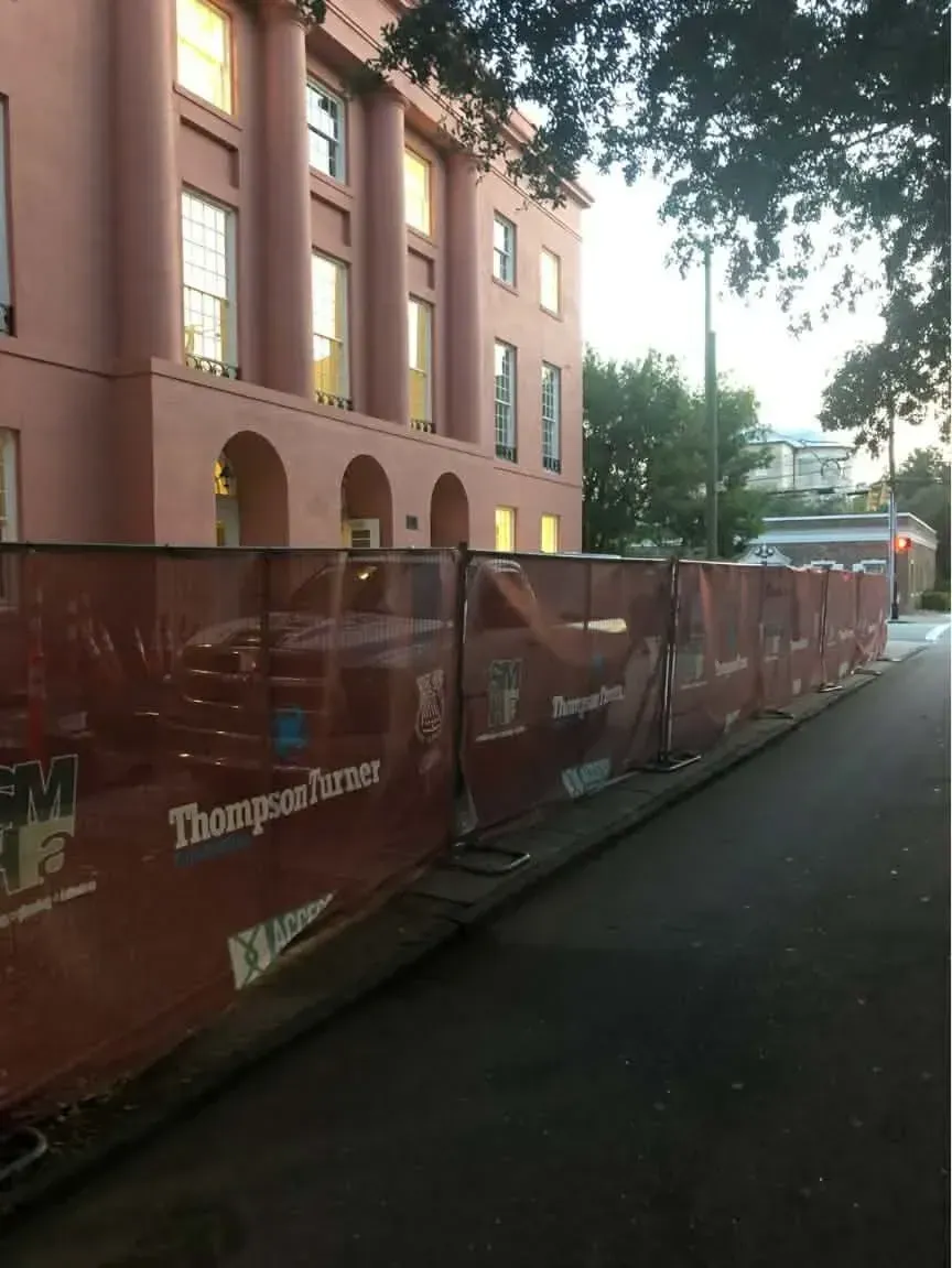 Red construction barrier along a wet street beside a brick building with trees overhead