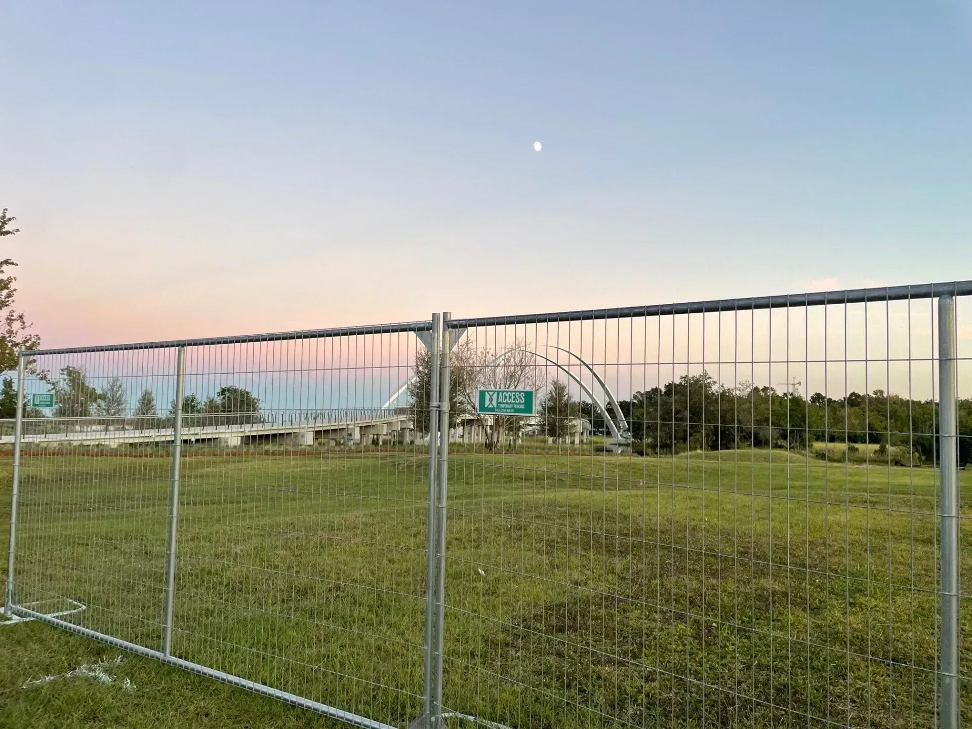 Chain-link fence in front of a grassy field at dusk, with a small sign and pastel sky.