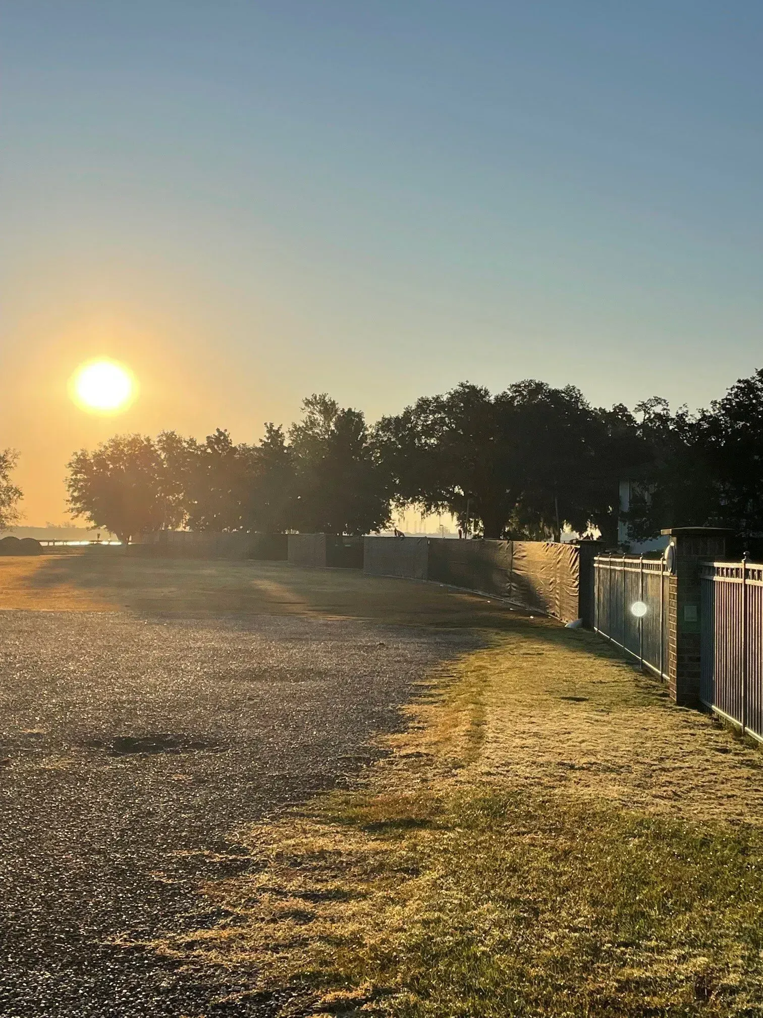 Sunrise over a gravel path beside fences and trees, with warm golden light across the ground.