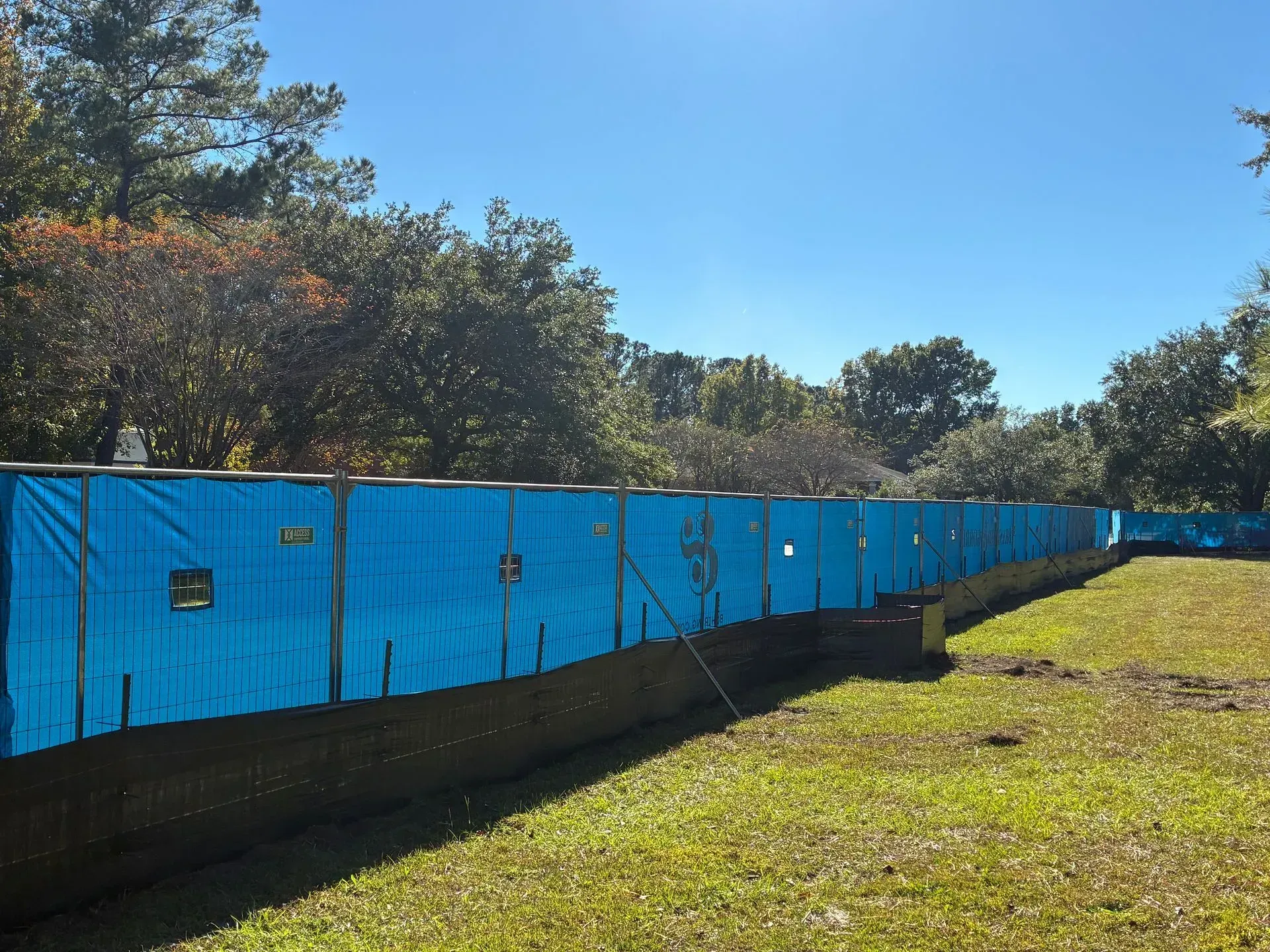 Blue construction fence along a grassy roadside under a clear sky, with trees in the background