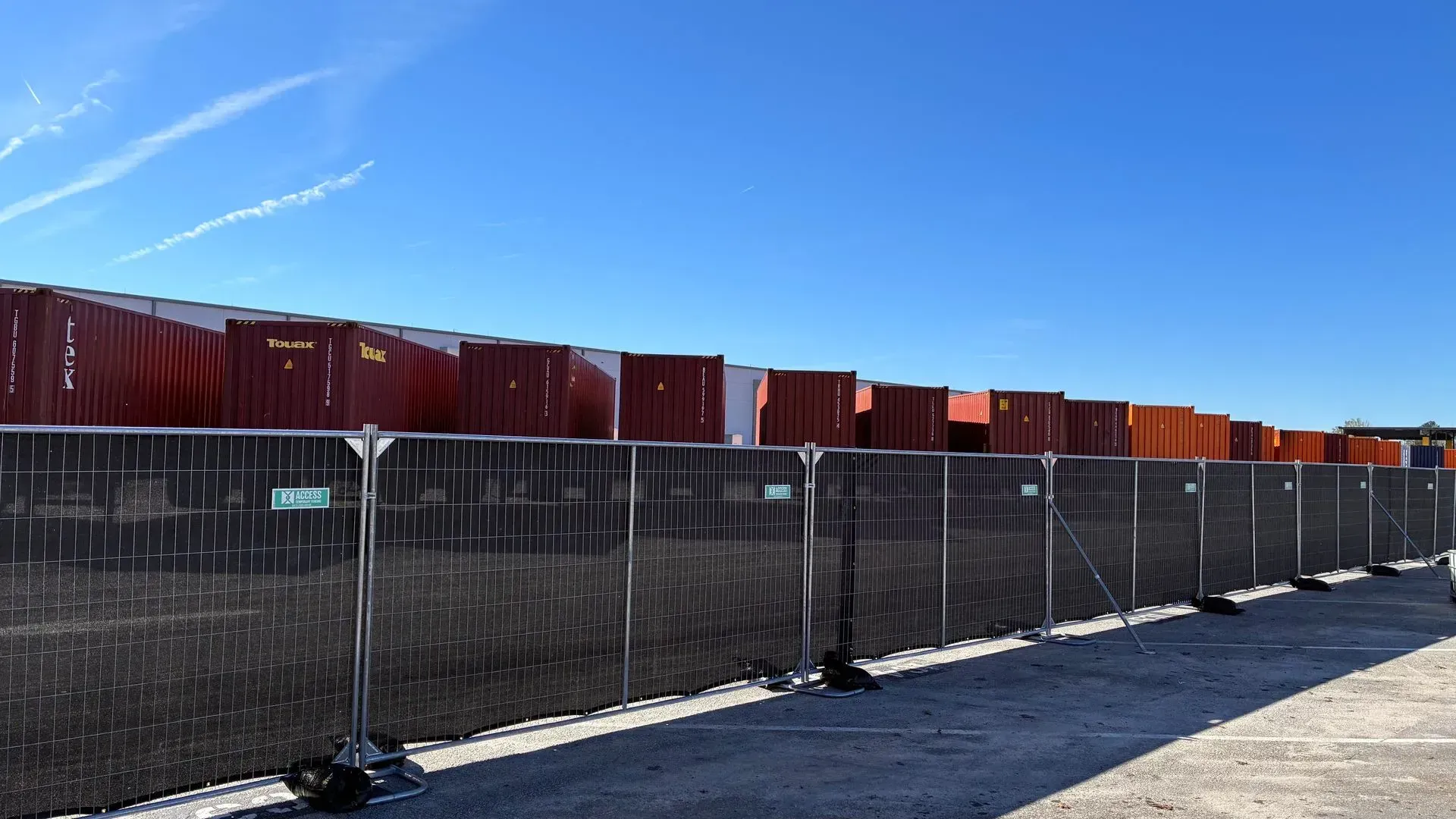 Row of stacked shipping containers behind a black fence under a bright blue sky
