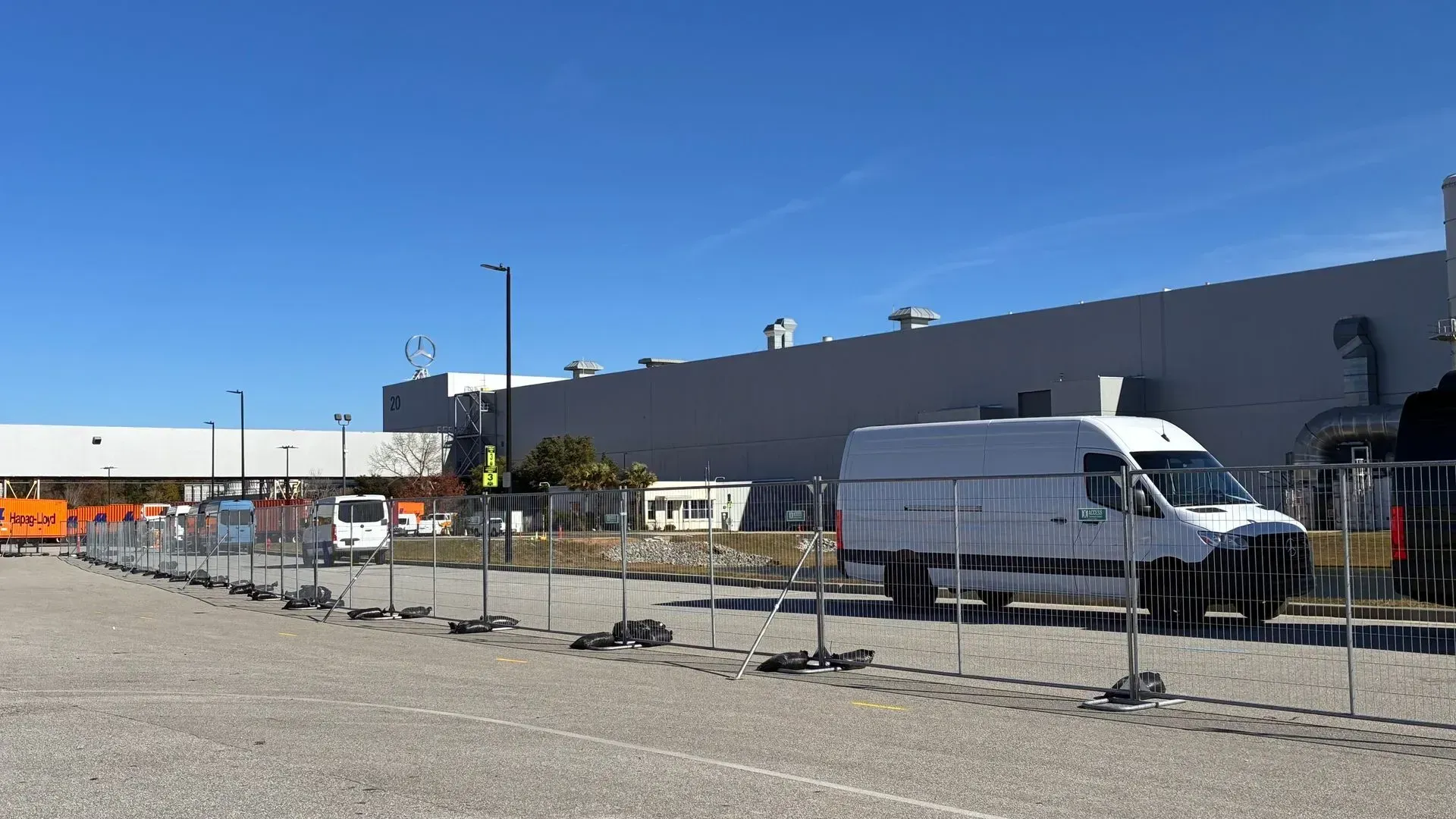 Parking lot beside a large gray industrial building, with white vans and metal fencing under a clear blue sky.