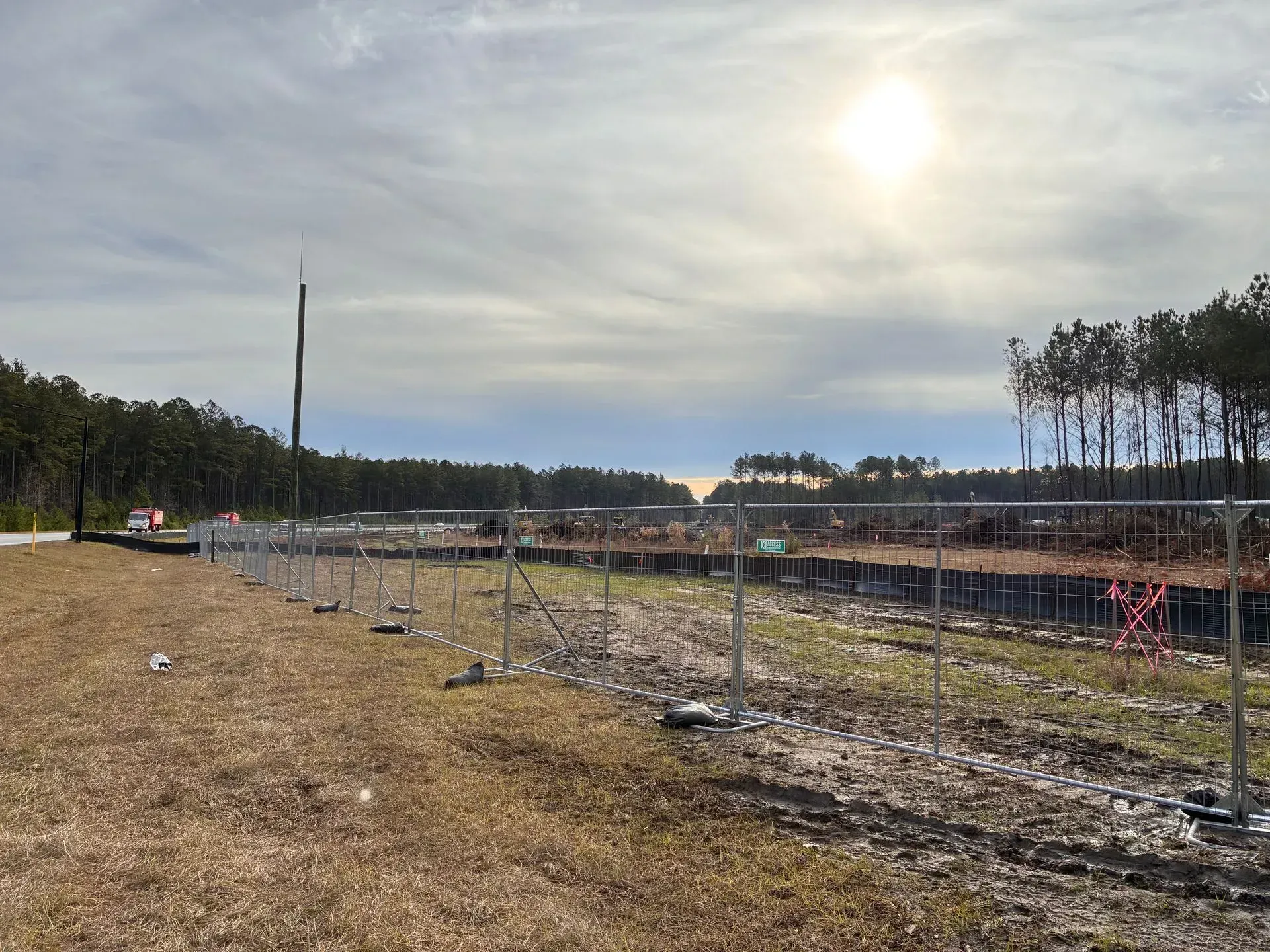 Rural construction site with fenced gravel lanes, sparse trees, and a bright cloudy sky