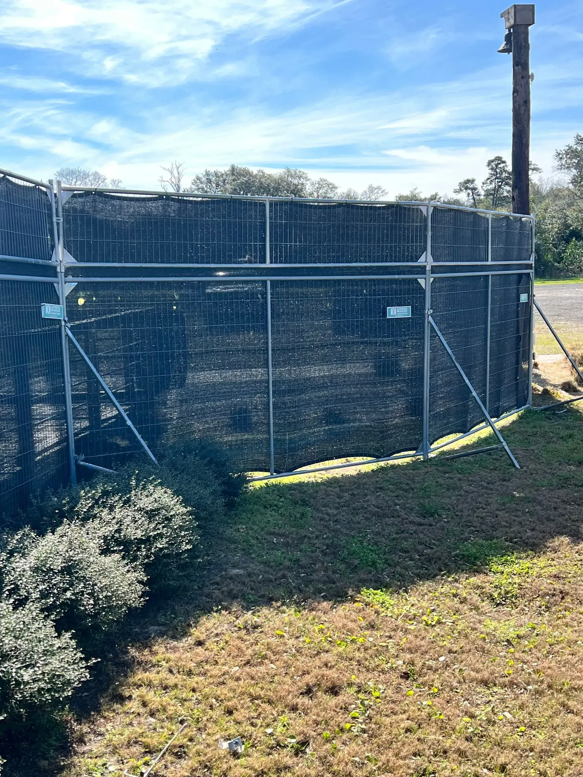 Chain-link fenced utility area with a tall pole, grassy foreground, and blue sky.