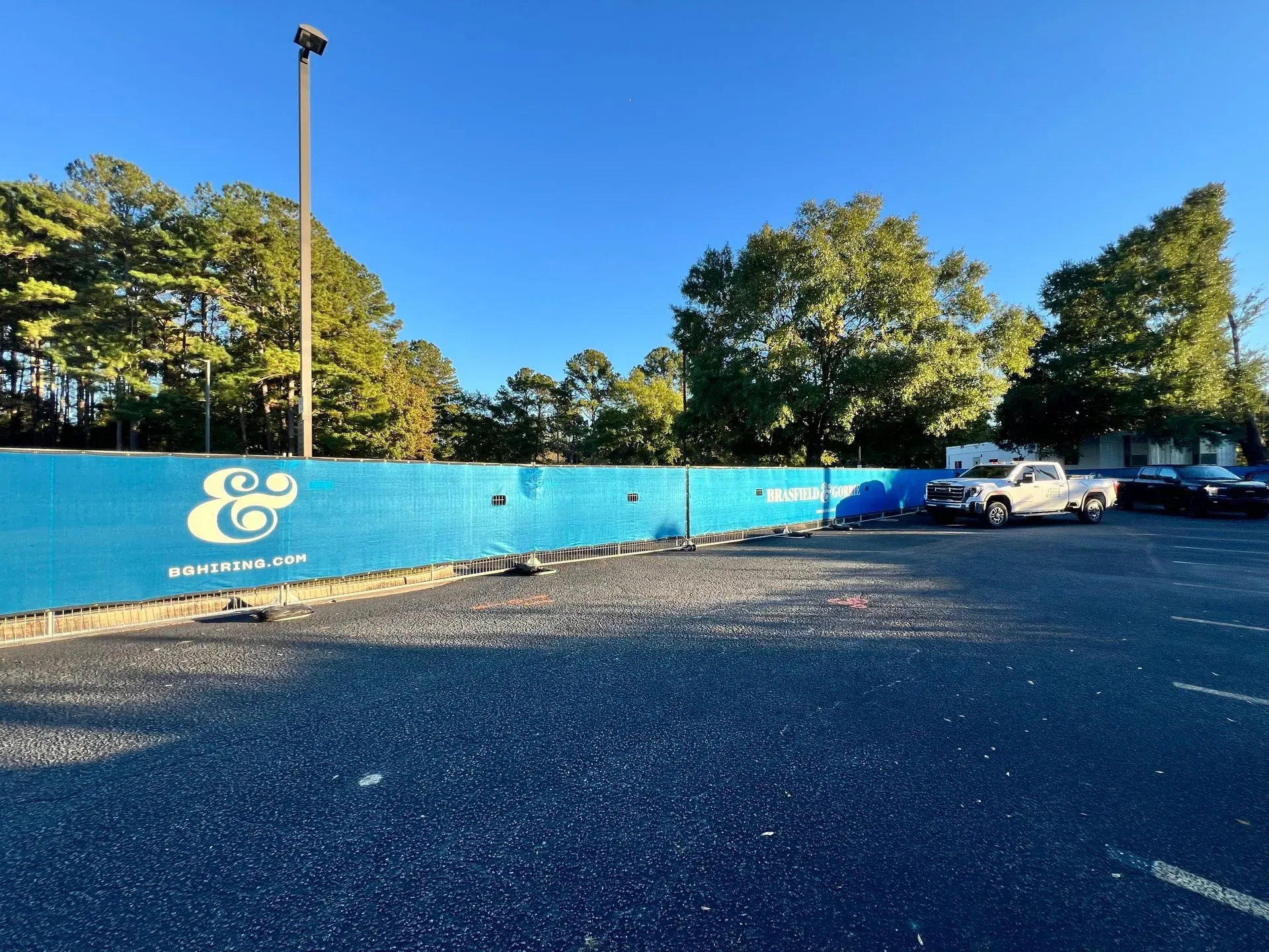 Blue wall with large white logo beside a gravel lot, parked cars, and trees under a clear sky