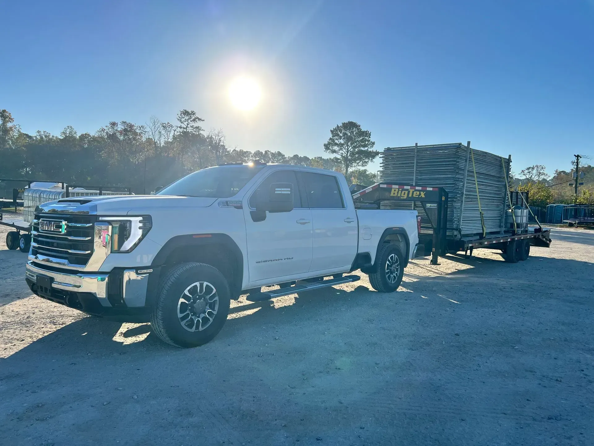 White pickup truck towing a metal trailer in a sunny gravel lot, with trees in the background
